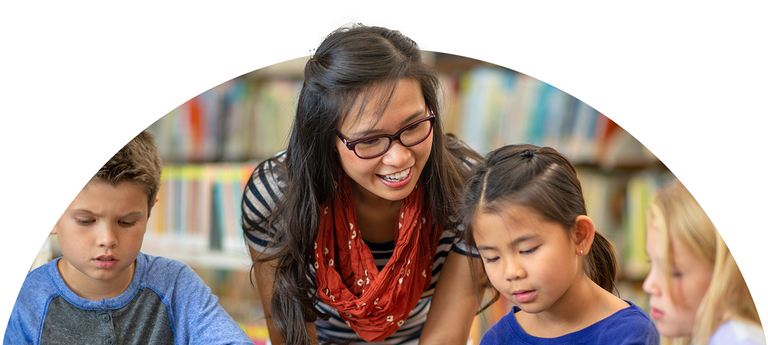 A female-presenting teacher - who has glasses and wears a red scarf - is kneeling over to talk to three seated children.
