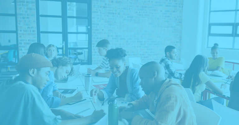 In a classroom, a group of ten teacher candidates sit around a large table. They write papers, collaborate with one another, and listen to what is being said.